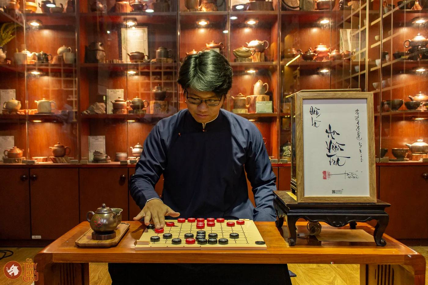 Hình ảnh Co-founder Cờ Thủy Tinh Châu chụp hình trước tủ trà, bàn Cờ Thủy Tinh Châu tại Đây là An Viên Trà - Trà Đạo